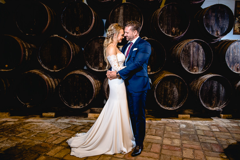 Pareja de recién casados besándose entre barricas de vino en la bodega de la hacienda Doña Caridad, fotografía por Moisés García para Bodas con Arte, fotógrafo de bodas en Sevilla Este.