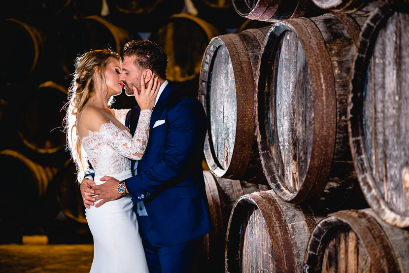 Pareja de recién casados besándose entre barricas de vino en la bodega de la hacienda Doña Caridad en el dia de su Boda en la Hermandad de los Negritos, fotografía por Moisés García para Bodas con Arte, fotógrafo de bodas en Sevilla Este.